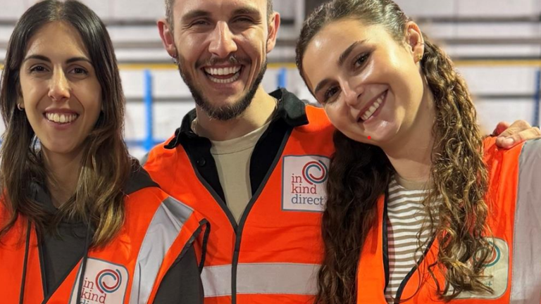 Three smiling volunteers wearing orange ""In Kind Direct"" vests standing together indoors."