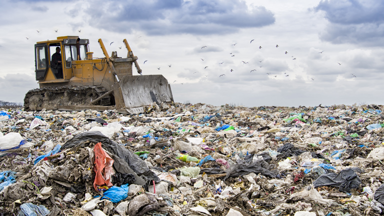 Bulldozer clearing garbage at a landfill with cloudy sky and birds in the distance.