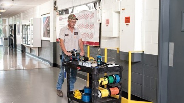 Maintenance worker pushing a tool cart with various equipment and cables through a hallway.