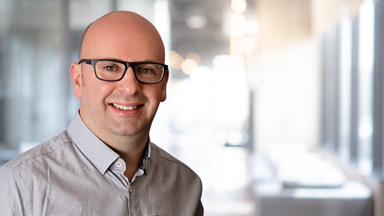 Bald man with glasses smiling in a blurred modern office setting.