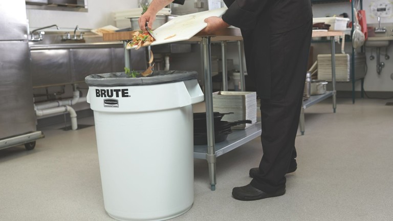 Chef discarding food waste into large white garbage bin in commercial kitchen.