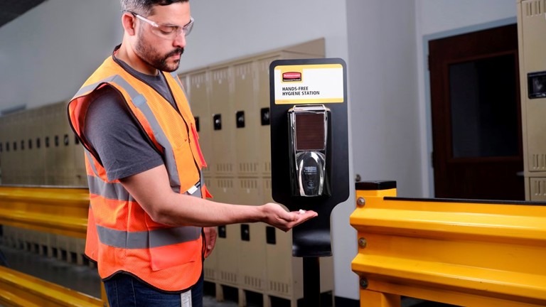 Worker in safety gear using hands-free hygiene station near lockers in industrial setting.