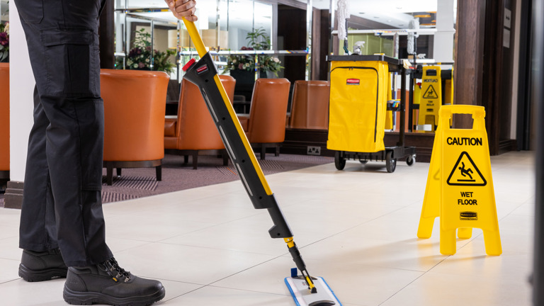 Janitor mopping floor in lobby with caution wet floor sign and cleaning cart nearby.