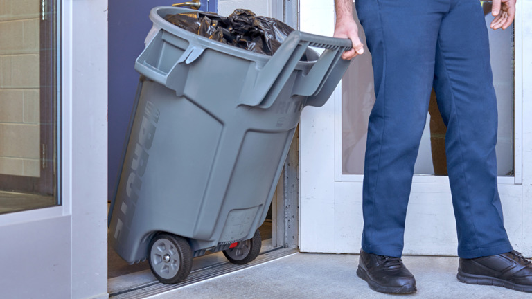 Person in blue pants wheeling a large gray trash can full of garbage bags through a doorway.