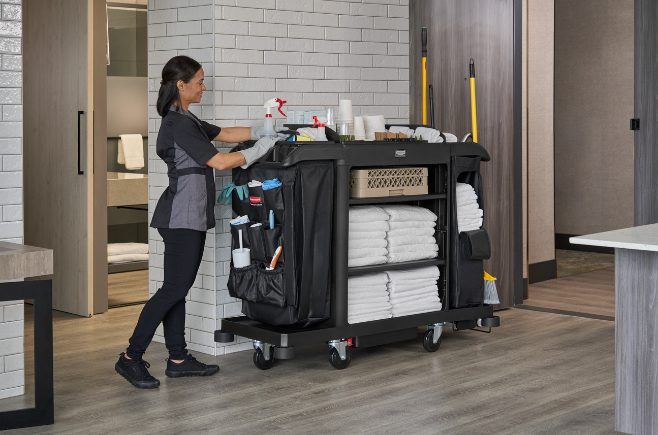 Housekeeping staff with a fully stocked cleaning cart in a modern hotel hallway.