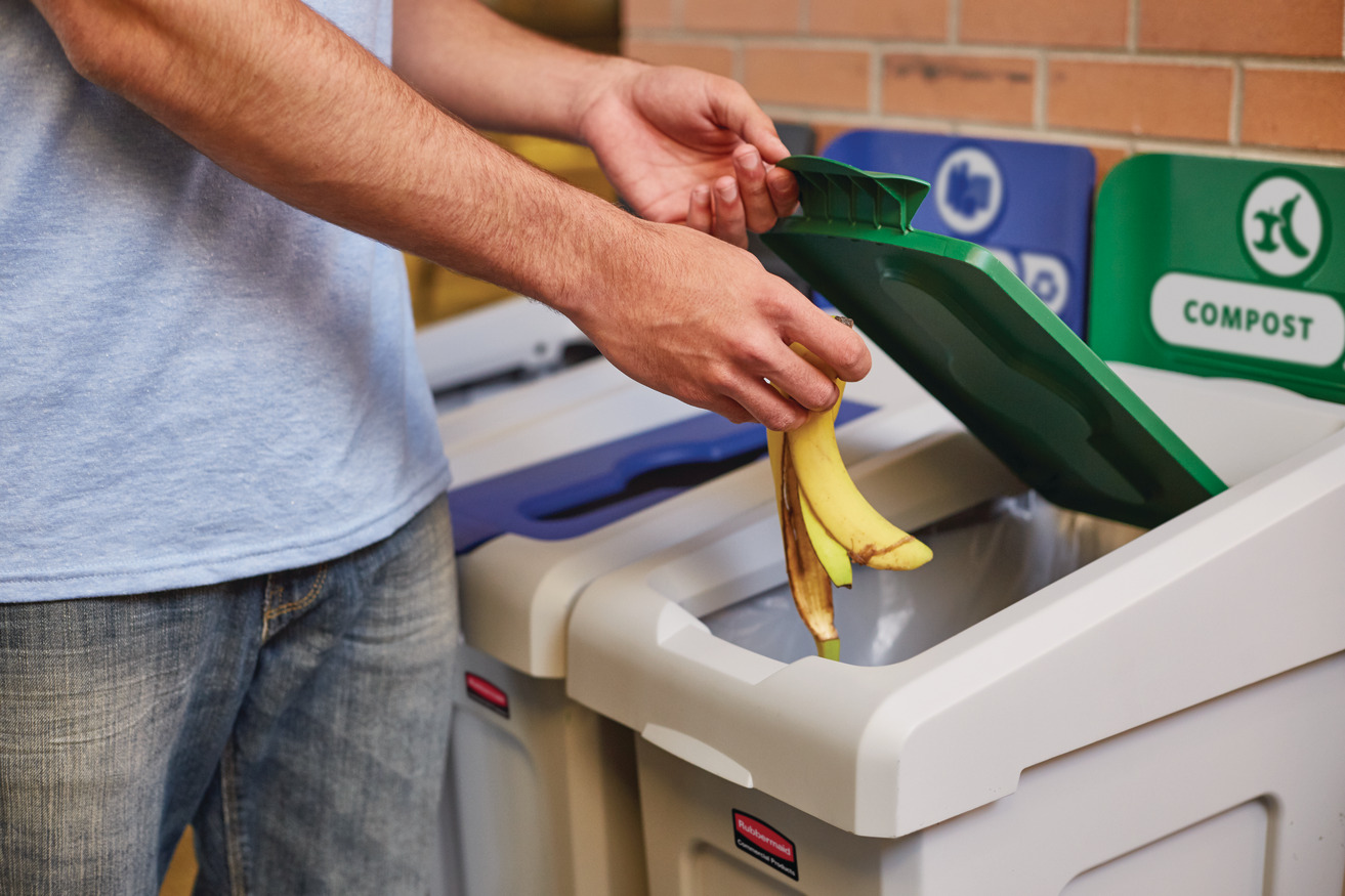Person composting banana peel in green-lid bin next to recycling containers