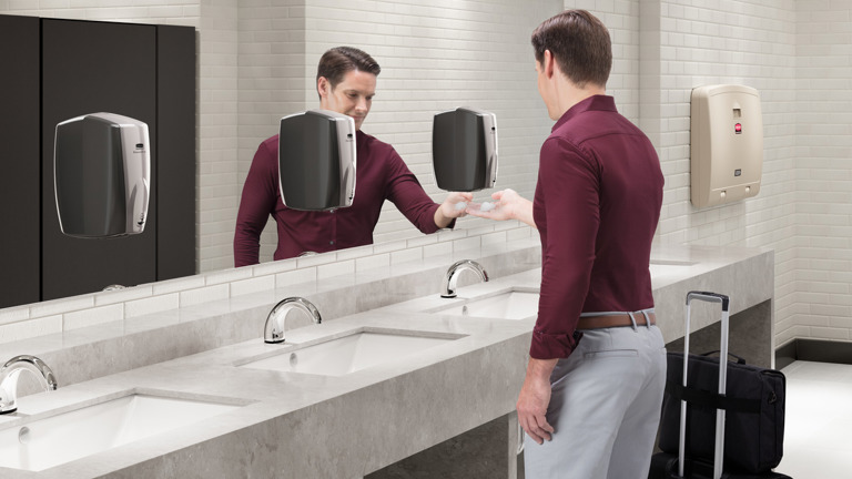 Man using a motion-activated soap dispenser in a modern restroom with a travel bag beside him.