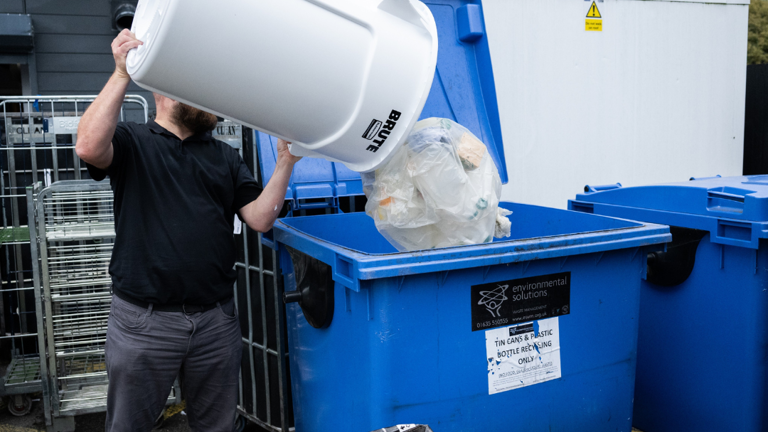 Person emptying white bin into large blue recycling dumpster outdoors.